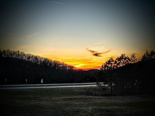 Sunset over a highway, with silhouettes of trees and rolling hills. The sky features vibrant orange and purple hues, creating a serene atmosphere.