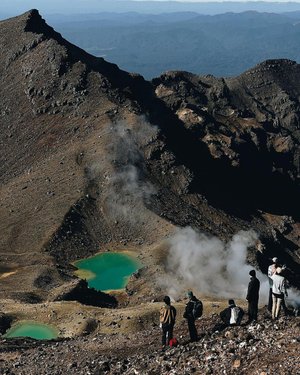 A group of hikers stands on a rocky mountain ridge, overlooking vibrant green lakes and steam rising from the geothermal landscape below.