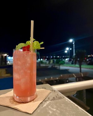 A refreshing pink cocktail garnished with mint and a raspberry sits on a coaster, with a softly lit outdoor setting in the background.