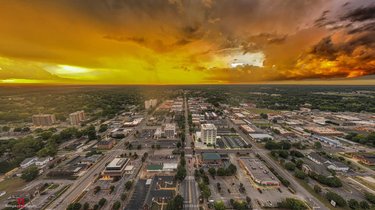 Aerial view of a vibrant cityscape at sunset, showcasing streets lined with buildings and greenery under a dramatic sky.