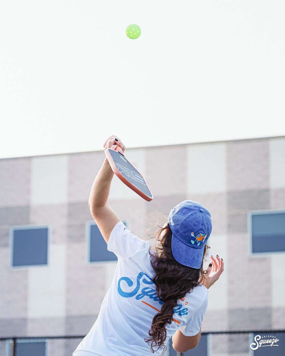 A person in a blue cap serves a green ball with a paddle in a sports court, surrounded by a modern building backdrop.
