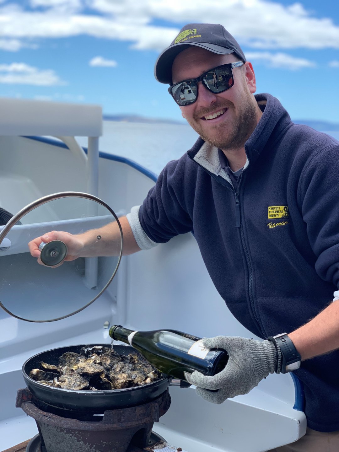 A man in a cap and sunglasses prepares oysters on a boat, holding a bottle of champagne. The ocean and sky are visible in the background.