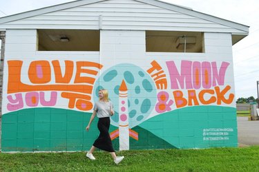 A woman walks past a colorful mural featuring the phrase "Love You to the Moon & Back," with a rocket and moon design. Green grass surrounds the scene.