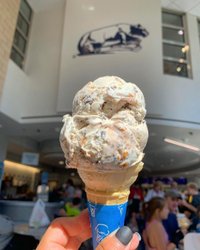 A hand holds a large ice cream cone with swirls of flavors, set against a bustling indoor space with people and a mural above.