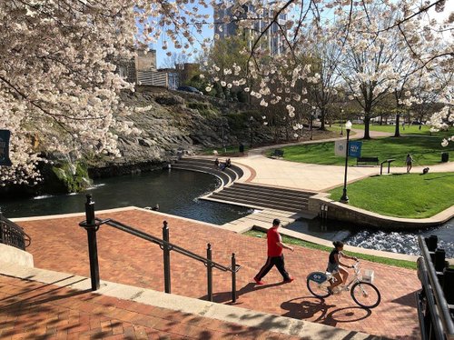 A scenic park with cherry blossom trees, a flowing stream, and pathways. A person walks while another rides a bike along the water's edge.