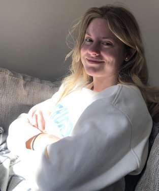 A young woman sits comfortably on a couch, smiling softly in natural light, wearing a cozy sweatshirt. The setting is warm and inviting.