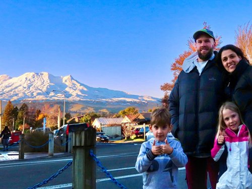 A family stands together in front of a snow-capped mountain, with trees and buildings in the background under a clear blue sky.