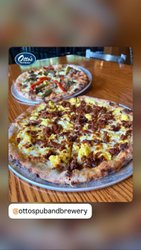 Two pizzas on metal plates sit on a wooden table. One features toppings of sausage and cheese, while the other has peppers.
