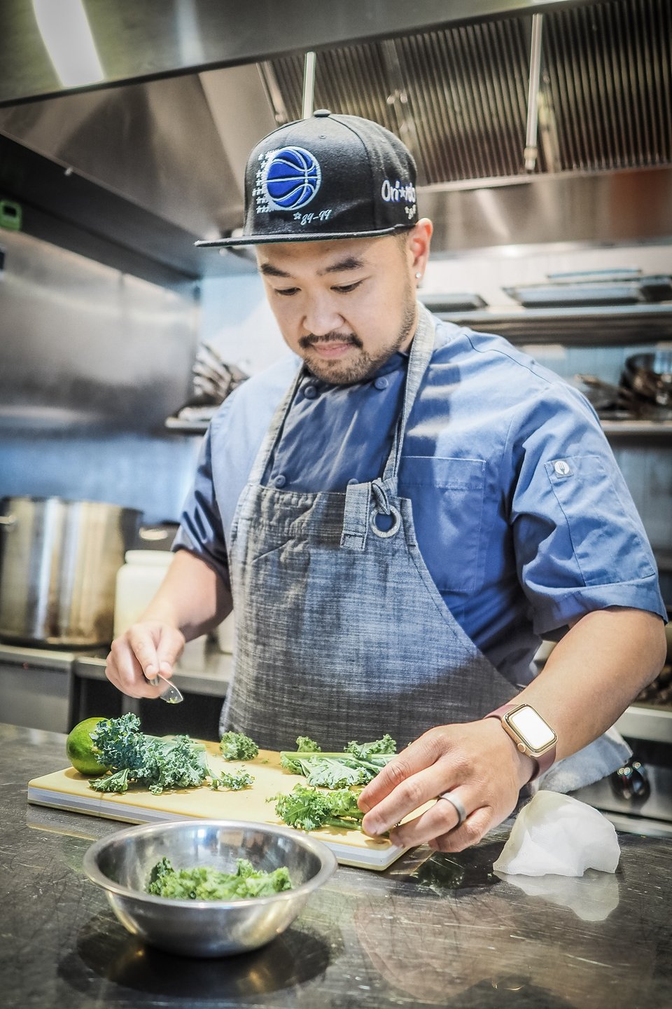 A chef in a modern kitchen prepares fresh kale on a cutting board, with a bowl of chopped greens nearby. Stainless steel appliances are visible.