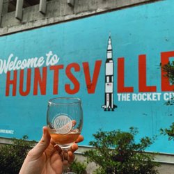 A hand holds a glass in front of a vibrant mural reading "Welcome to Huntsville, The Rocket City," featuring a rocket illustration and greenery.