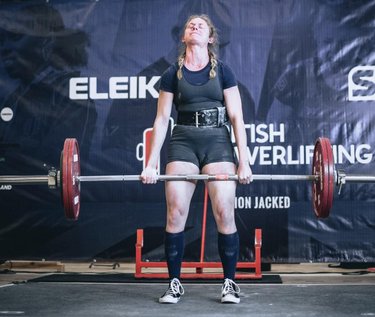 A female weightlifter performs a deadlift with a barbell, set against a backdrop featuring the ELEIKO logo and competition branding.