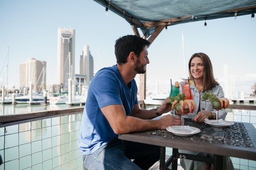 A couple enjoys colorful cocktails and seafood dishes at a waterfront table, with a marina and city skyline in the background.