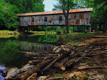 A rustic covered bridge spans a calm, reflective stream, surrounded by lush greenery and rocky banks, inviting exploration and tranquility.