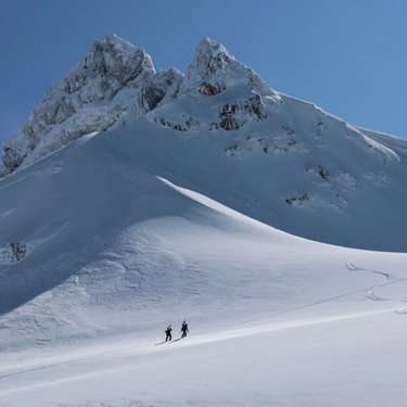 Two hikers traverse a vast, snowy landscape beneath towering, jagged mountains. The clear blue sky enhances the serene, remote atmosphere.