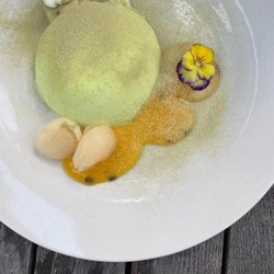 A round green dessert sits on a white plate, accompanied by two scoops of sorbet, a vibrant sauce, and a purple flower garnish.