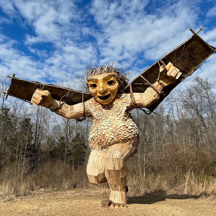 A large wooden sculpture of a mythical creature with outstretched wings stands in a natural setting,…