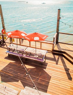Wooden deck overlooking calm water, featuring two red umbrellas shading picnic tables. Soft sunlight reflects on the surface.