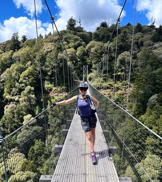 A person stands confidently on a suspension bridge surrounded by lush green trees and a bright blue sky, showcasing an adventurous outdoor experience.