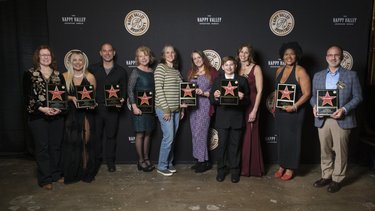 Group of ten individuals standing in front of a black backdrop with a logo, holding awards with red stars. The setting appears celebratory.