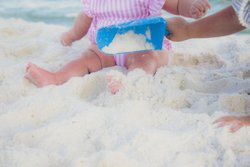 A child in a pink striped outfit sits on soft white sand, playing with a blue shovel. Another hand reaches in to assist.