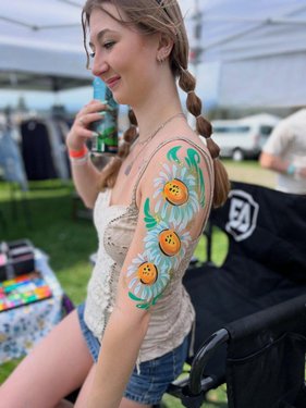 A young woman with braided hair shows off a colorful floral arm tattoo while sitting in a chair at an outdoor event. Green grass and tents are visible in the background.