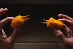 Two hands hold two golden, breaded crab claws, showcasing their crispy texture and visible crab pincers against a dark background.