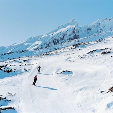 Two skiers navigate a winding snow-covered slope, surrounded by rugged mountains under a clear blue sky. The scene captures a winter landscape ideal for outdoor adventures.