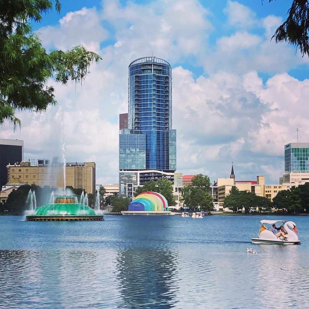A vibrant cityscape featuring a lake with colorful fountains, paddle boats, and modern skyscrapers under a partly cloudy sky.