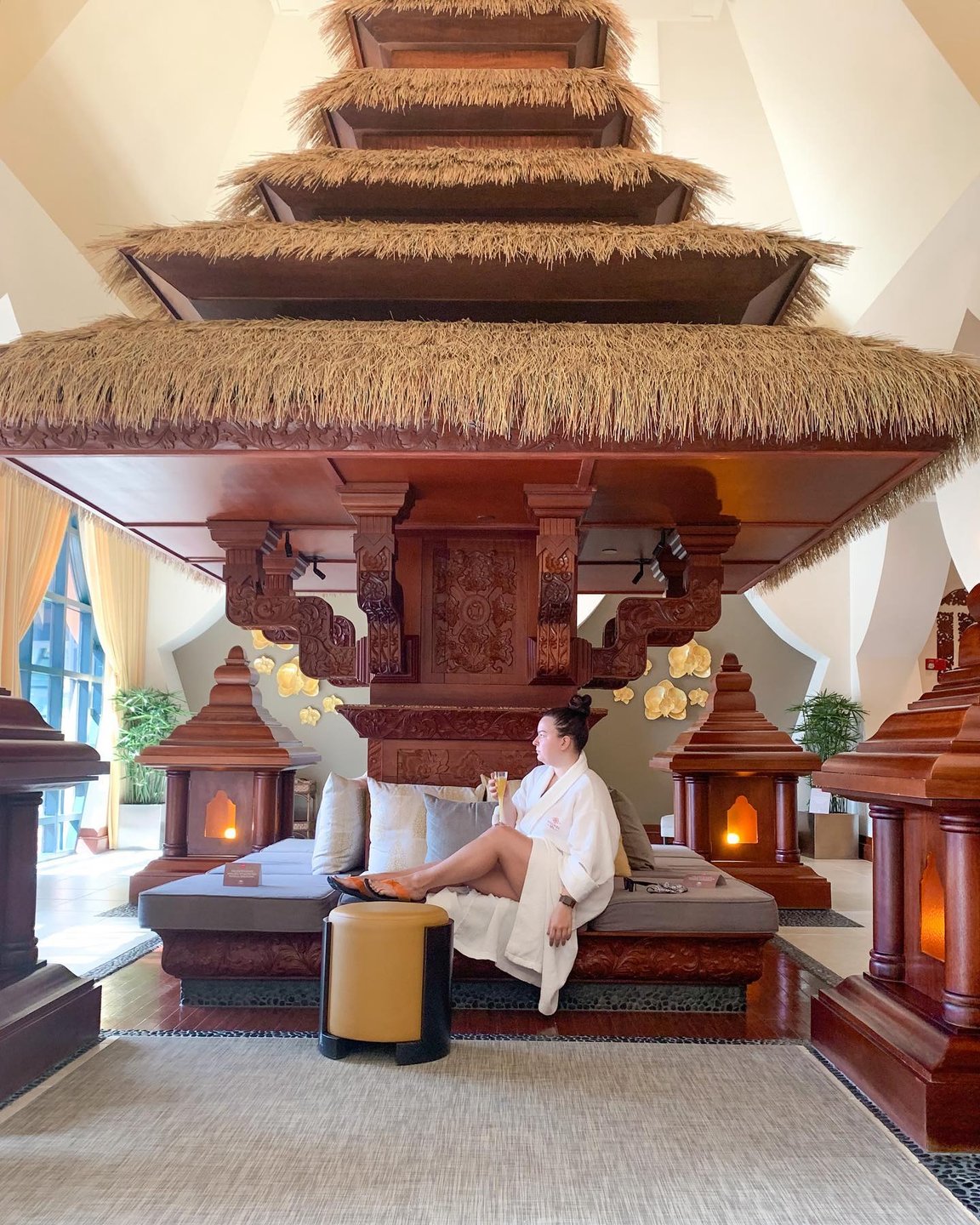 A serene spa setting featuring a woman in a robe relaxing under a large, intricately carved wooden structure with soft lighting.