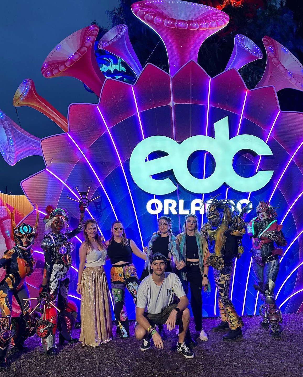 A vibrant scene at a festival with a large illuminated shell backdrop displaying "EDC Orlando." Six people pose with colorful performers in costumes.