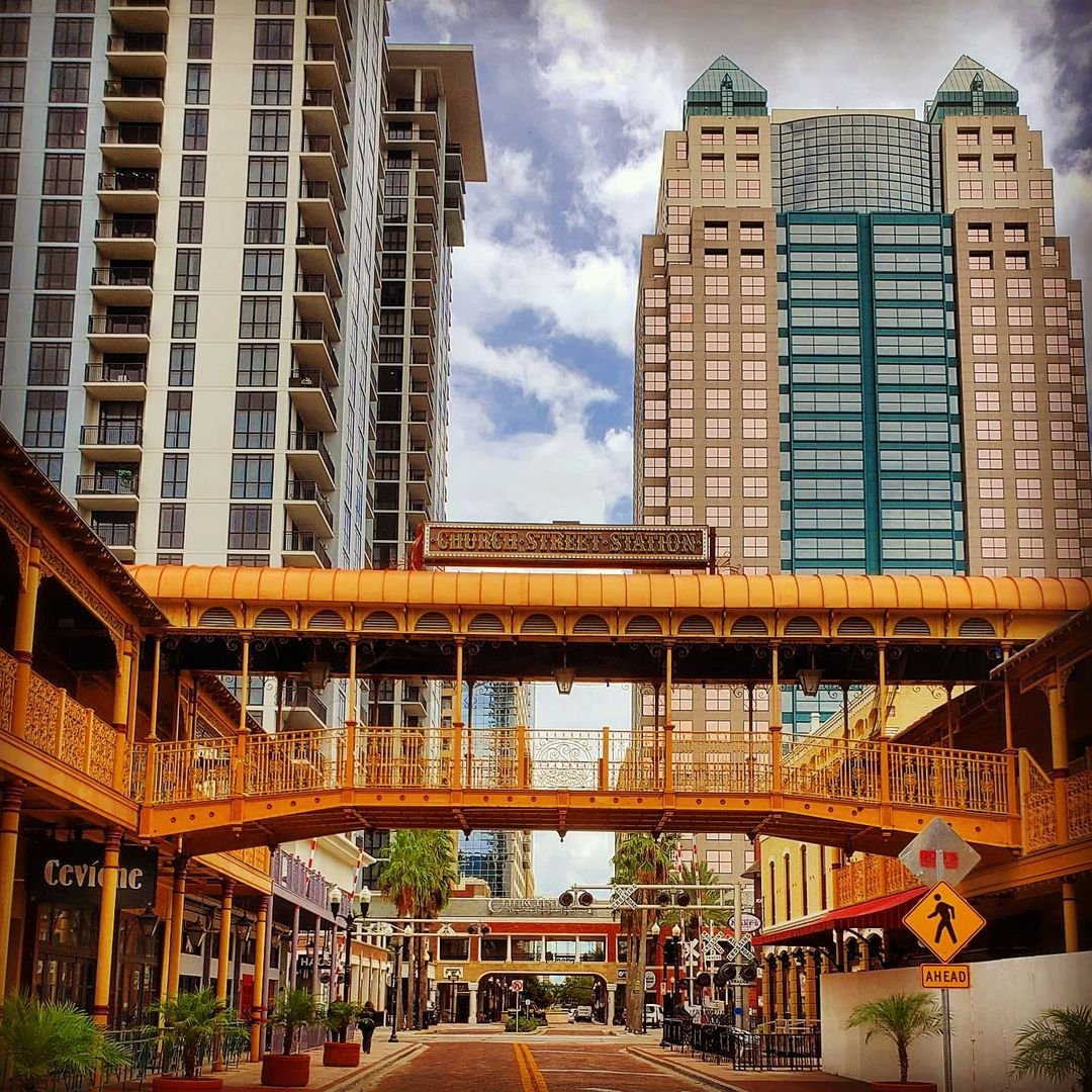 A vibrant urban street scene featuring a historic elevated walkway connecting buildings, flanked by modern skyscrapers and shops below.