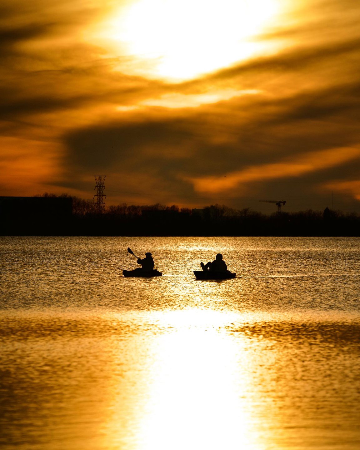 Two kayakers paddle across a shimmering lake at sunset, with vibrant clouds reflecting on the water'…