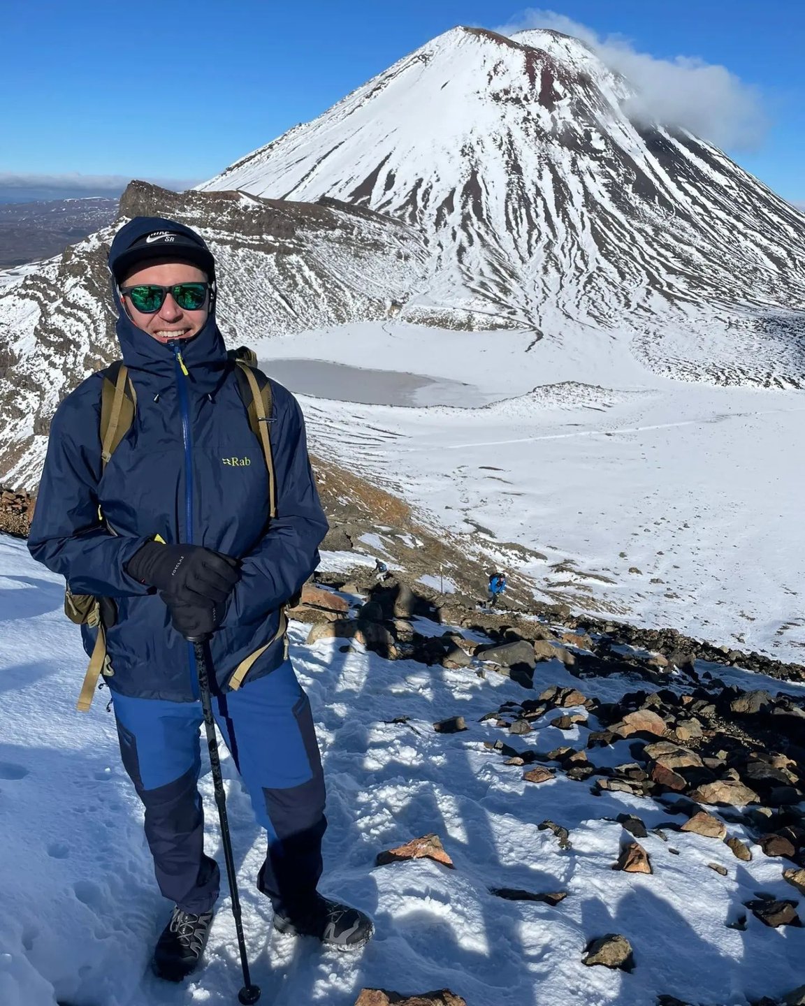 A hiker stands on a rocky slope, wearing winter gear, with a snow-capped volcano in the background under a clear blue sky.