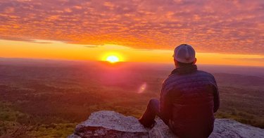 A person sits on a rocky ledge, gazing at a vibrant sunset over a vast landscape of rolling hills and valleys. The sky is filled with colorful clouds.