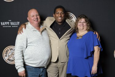 Three individuals pose together in front of a dark backdrop featuring the "Happy Valley Adventure Bureau" logo. They smile, dressed in casual and semi-formal attire.