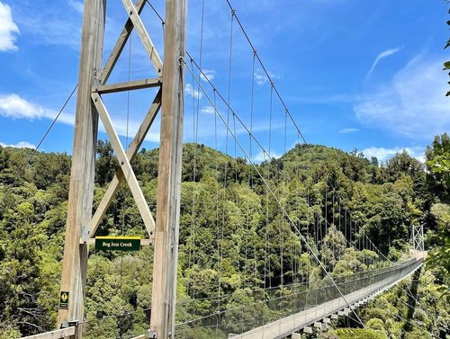 Suspension bridge spans a lush green valley, surrounded by dense trees and hills under a clear blue sky. Sign reads "Big Iron Creek."