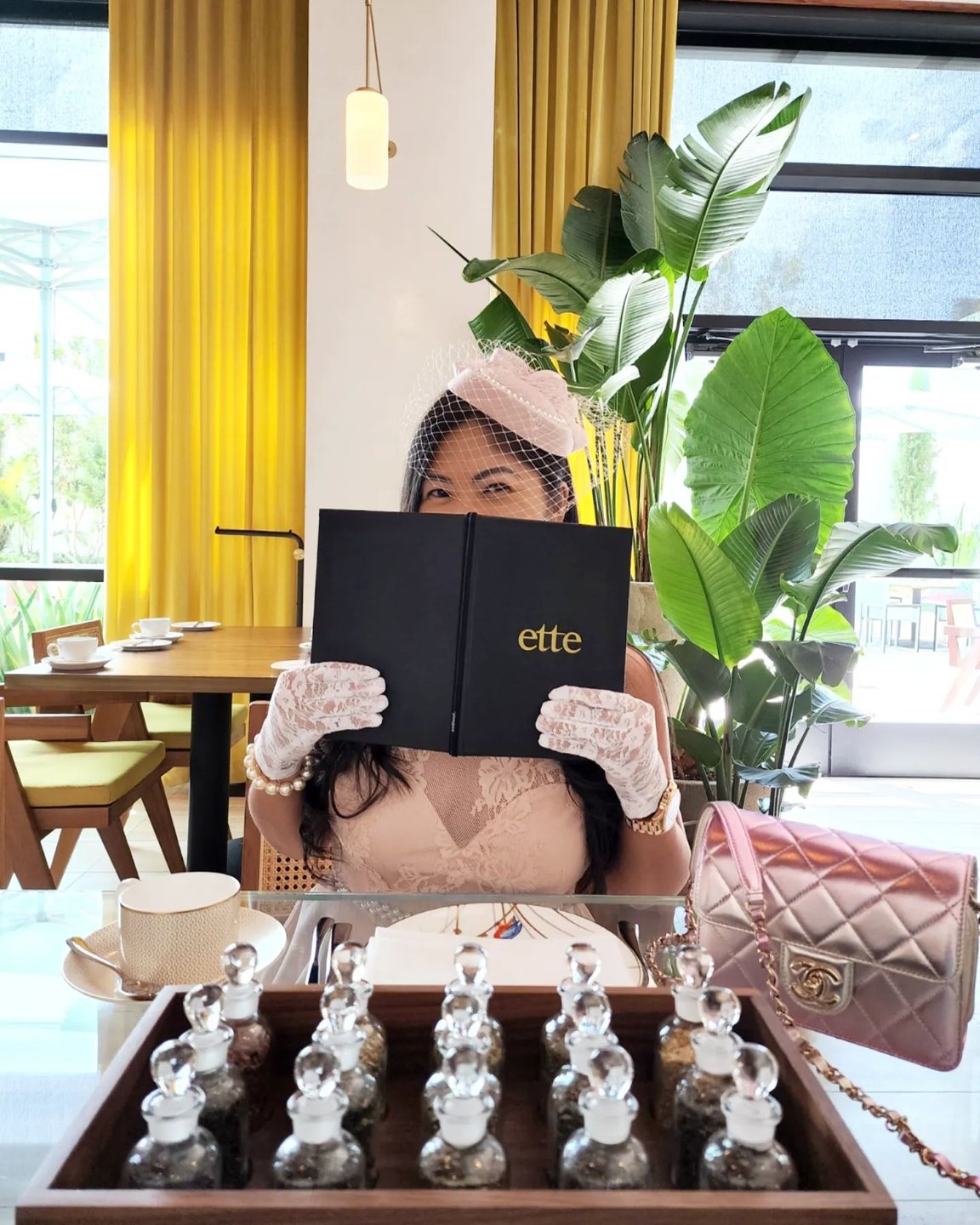 A woman in elegant attire holds a menu at a stylish restaurant, surrounded by greenery and modern decor. A tray of glass jars is in front of her.