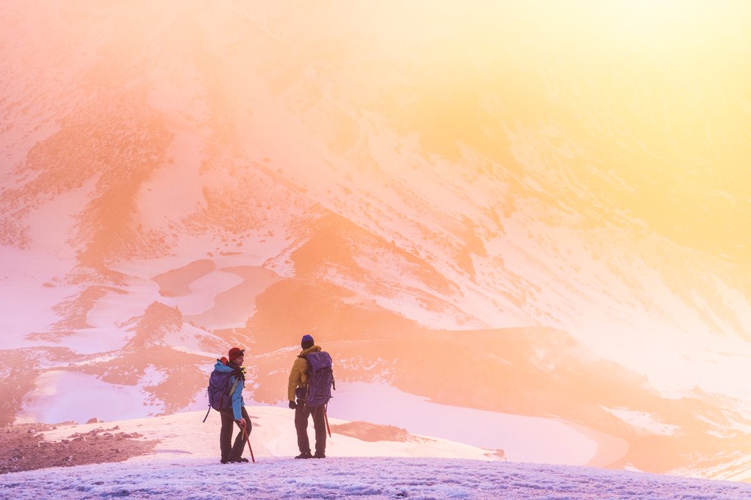 Two hikers stand on a snowy mountain ridge, overlooking a vast landscape bathed in soft, warm light. The scene captures a serene, adventurous atmosphere.