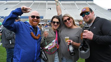 Four friends celebrate at an outdoor event, smiling and holding drinks. A stadium backdrop and festive atmosphere enhance the scene.