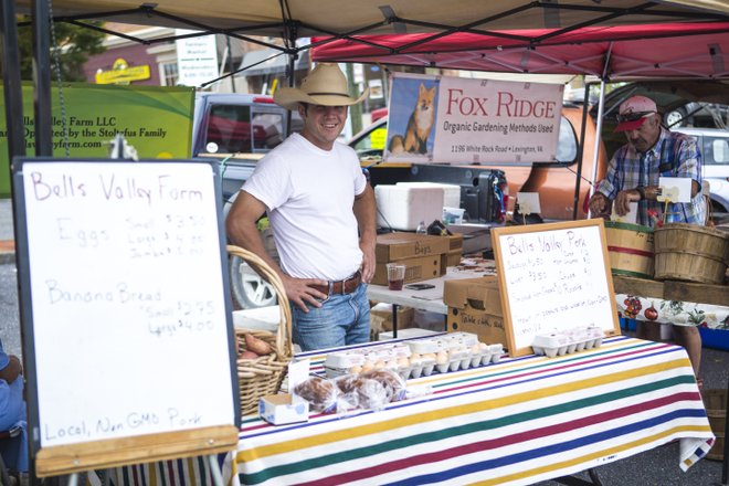 A farmer stands at a market stall with signs listing local products like eggs and banana bread. Baskets and goods are displayed under a colorful canopy.
