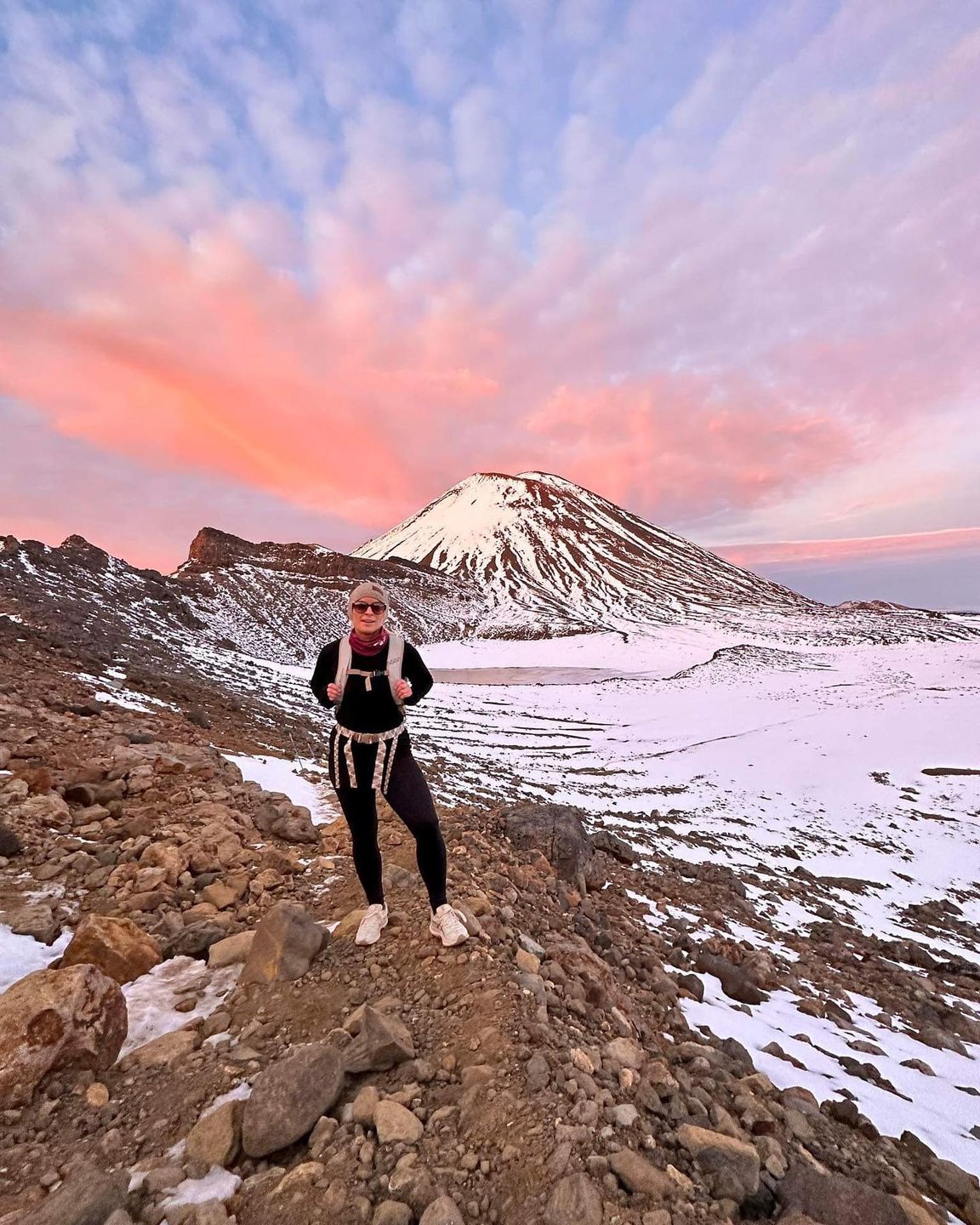 A hiker stands on a rocky path with a snow-covered mountain in the background, under a colorful sunset sky. The scene captures a serene, adventurous landscape.