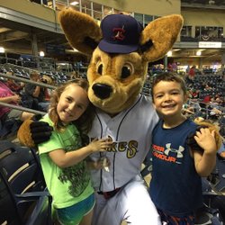 Two children pose with a large, friendly dog mascot in a sports stadium. The background shows empty seats and a lively atmosphere.