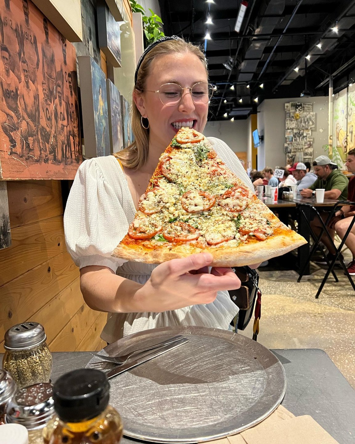 A woman holds a large slice of pizza topped with herbs and cheese, smiling in a bustling restaurant with patrons in the background.