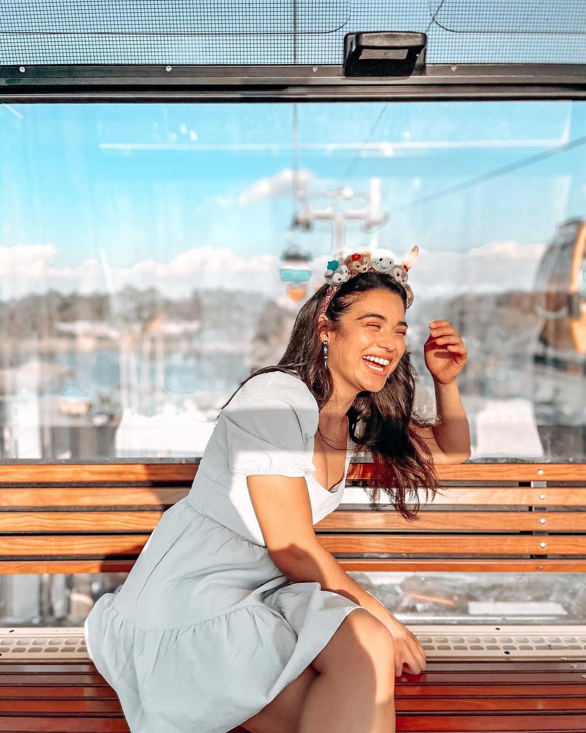 A woman in a light dress smiles while sitting on a wooden bench inside a gondola, with scenic views of mountains and a cable car in the background.