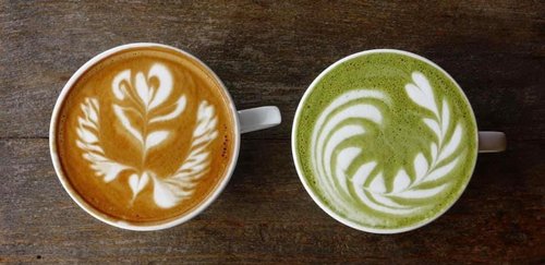 Two cups of artfully crafted beverages sit on a wooden surface: a latte with intricate white foam designs and a matcha drink with similar patterns.