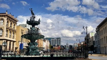 A historic fountain stands in a bustling city square, surrounded by charming buildings and a clear blue sky with scattered clouds.