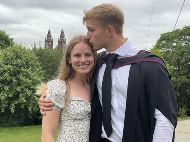 A young man in graduation attire kisses the forehead of a smiling woman outdoors, with historic buildings and greenery in the background.