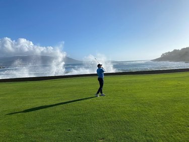 A golfer swings on a lush green course by the ocean, with waves crashing against the shore and mountains in the background.