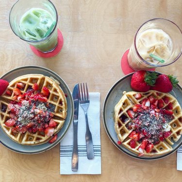 Two plates of waffles topped with fresh strawberries and chia seeds, accompanied by iced green tea and iced coffee, on a wooden table.