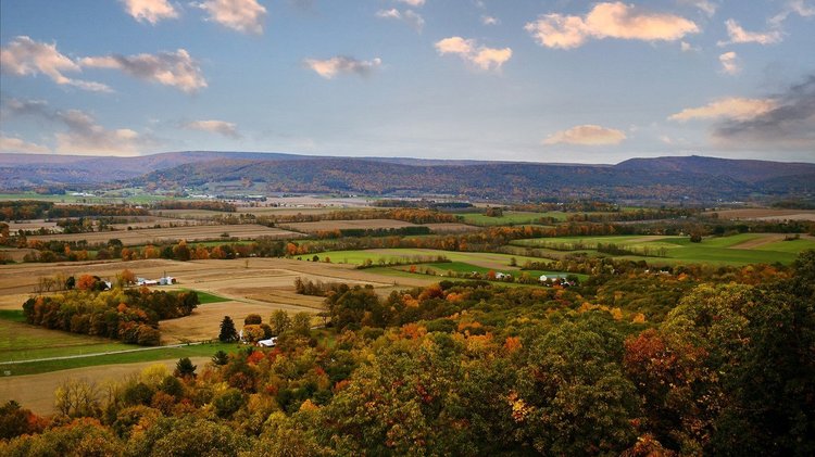 Vast landscape featuring rolling hills, vibrant autumn foliage, and farmland under a partly cloudy sky. Scenic view invites exploration.